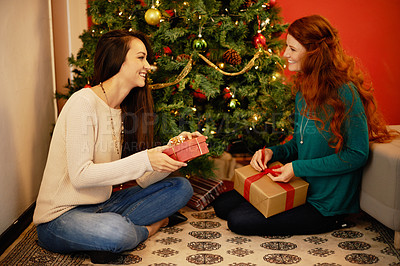 Buy stock photo Shot of two young women sitting by a Christmas tree and opening their presents
