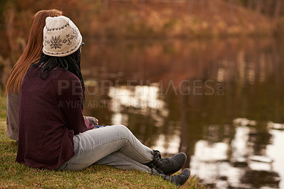 Buy stock photo Shot of two teenage girls sitting by a lake in the early evening