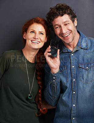 Buy stock photo Happy couple, phone and listening with call or voice note for communication or funny joke on a dark studio background. Man and woman with smile on mobile smartphone for online conversation or chat