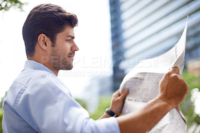 Buy stock photo A handsome young businessman reading a newspaper while sitting on a bench in the city