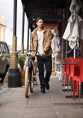 Buy stock photo Shot of a fashionable young man pushing his bicycle on the sidewalk