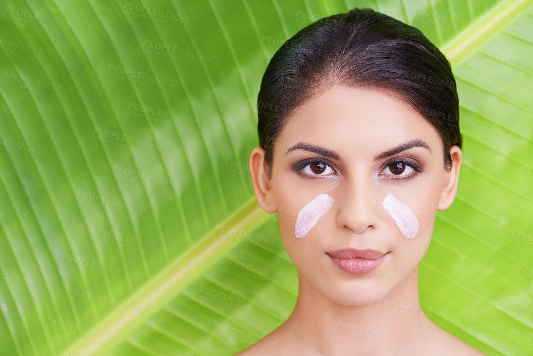 Buy stock photo Studio portrait of a beautiful young woman with moisturizer on her face posing in front of a leaf