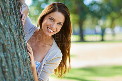 Buy stock photo Shot of a beautiful young woman standing behind a tree at the park