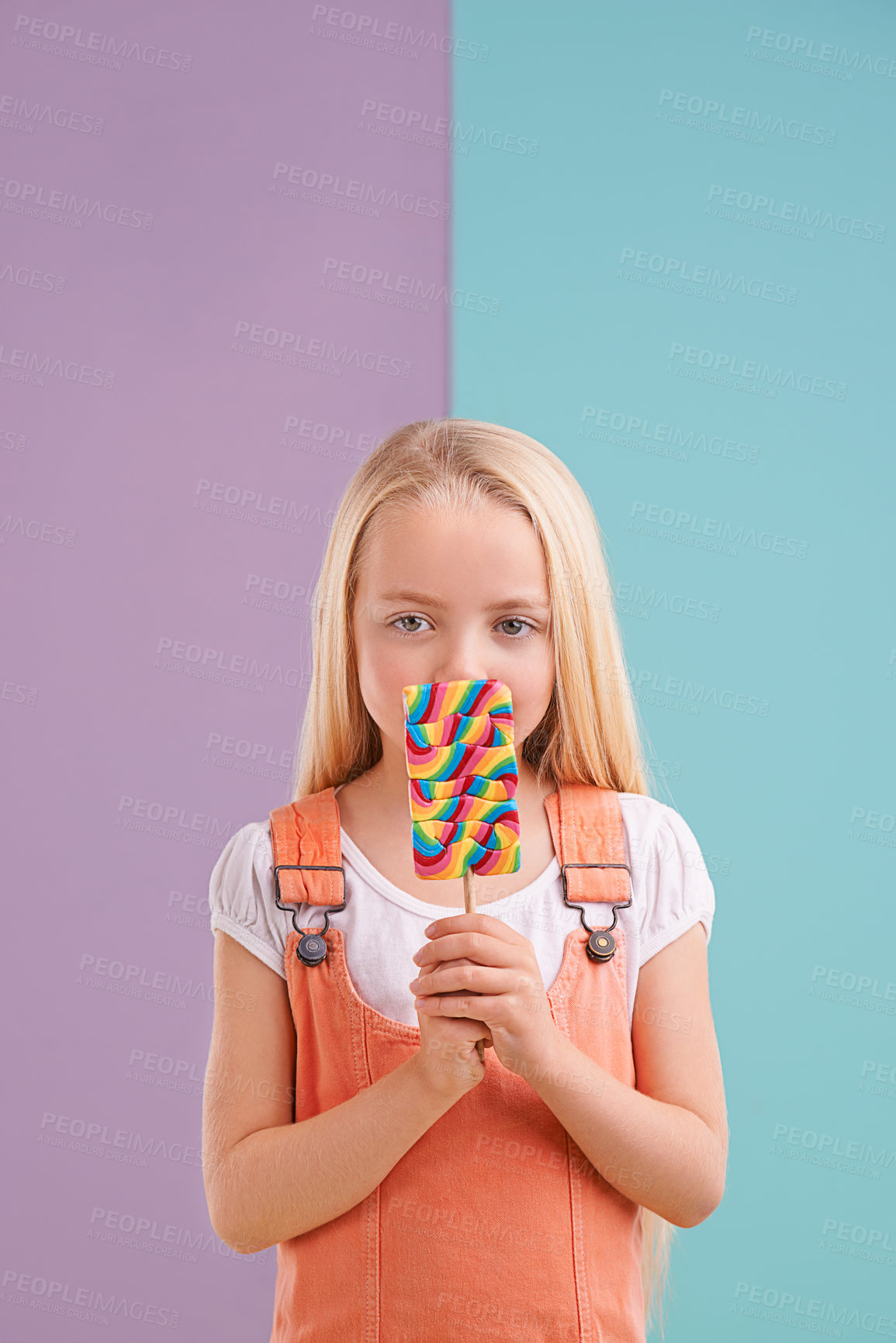 Buy stock photo Studio, child and hiding with lollipop, desert and playful for snack and childhood. Girl, sweets and yummy candy for tasty, eating and happiness on vibrant split pastel pink and blue background