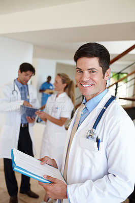 Buy stock photo Portrait of a male doctor standing with medical records with his colleagues in the background
