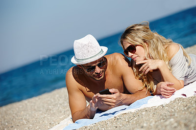Buy stock photo Shot of a young couple using a cellphone while suntanning on a beach