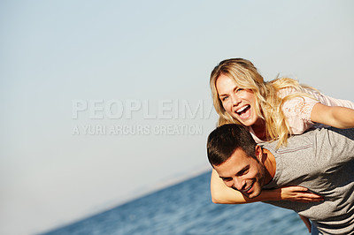 Buy stock photo Shot of a handsome young man giving his laughing girlfriend a piggyback at the beach