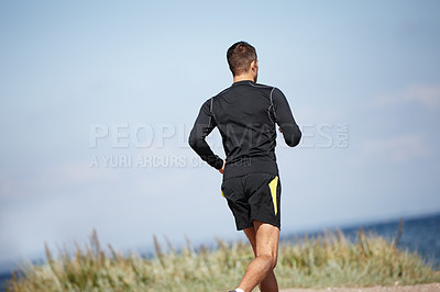 Buy stock photo Man, running and blue sky at beach for cardio with fitness motivation, exercise and health workout. Male athlete, training and nature by ocean with grass for wellness, strong body and endurance