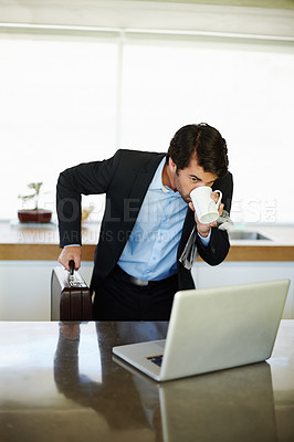 Buy stock photo Shot of a rushed businessman drinking coffee and using his laptop in his kitchen 