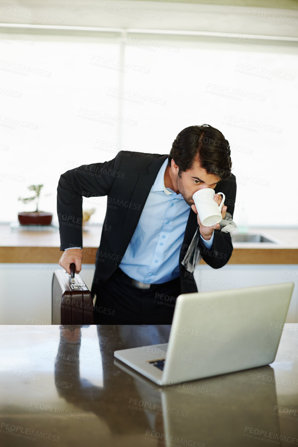 Buy stock photo Shot of a rushed businessman drinking coffee and using his laptop in his kitchen 