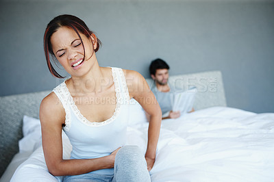 Buy stock photo Shot of a woman sitting in her bed with bad stomach pains with her boyfriend in the background