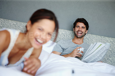 Buy stock photo Shot of a man in bed smiling at his girlfriend with a newspaper and mug in his hand