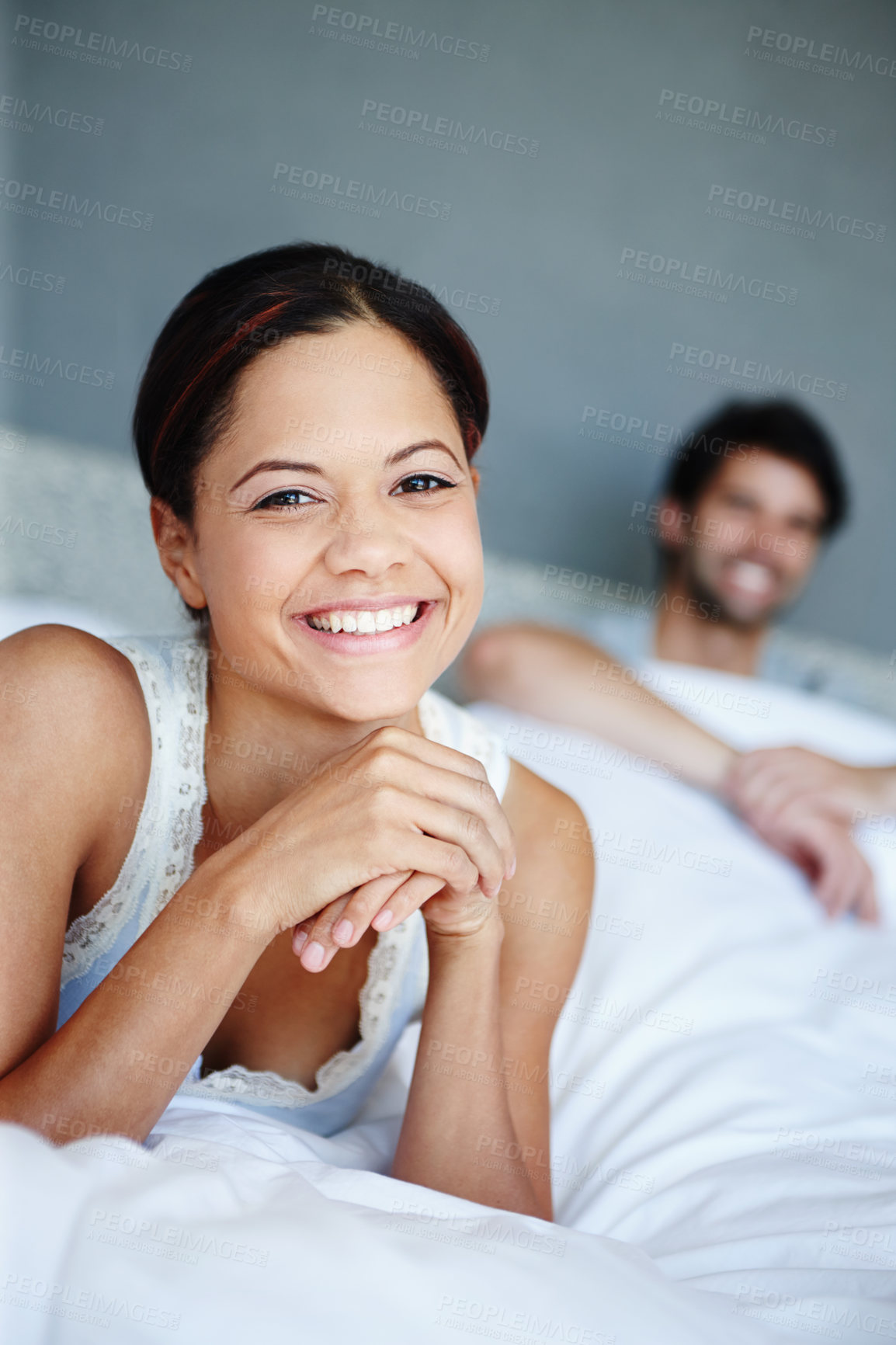 Buy stock photo Portrait of a smiling woman lying on her bed with her boyfriend in the background