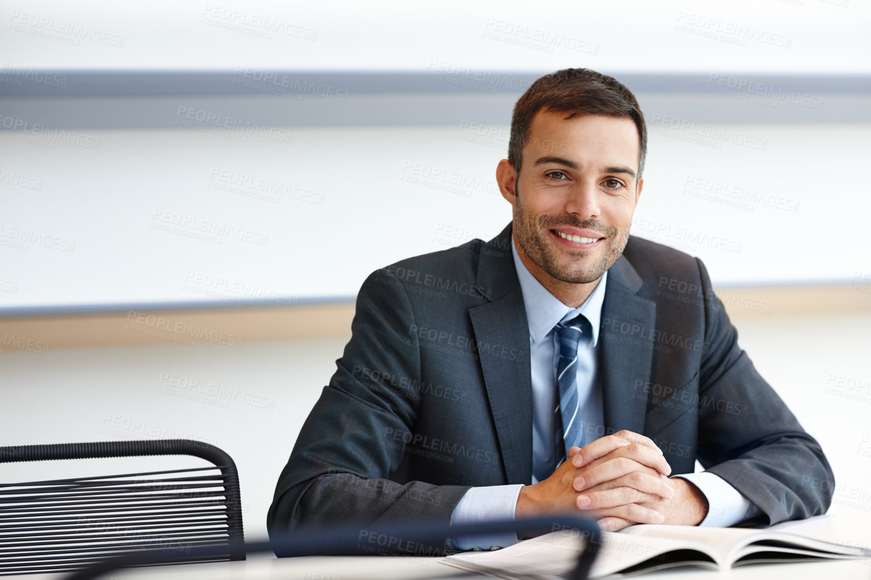 Buy stock photo Portrait of a young businessman sitting behind a desk in a bright office. Confident male business professional reading a magazine and wearing a suit. Proud boss smiling at work researching