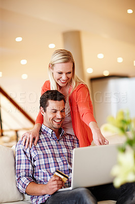 Buy stock photo A handsome man sitting on the couch with his laptop holding a credit card while his wife stands behind him