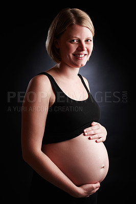 Buy stock photo Portrait of a pregnant woman holding her stomach isolated with a black studio background. Happy and smiling female expecting a baby standing alone inside a room. Attractive mom with an unborn child