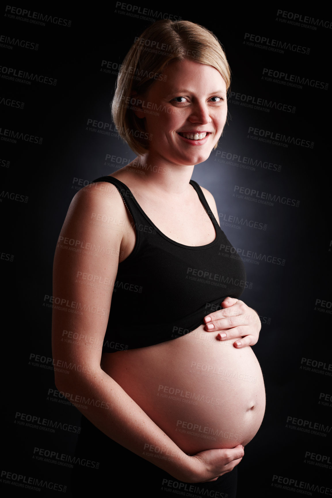 Buy stock photo Portrait of a pregnant woman holding her stomach isolated with a black studio background. Happy and smiling female expecting a baby standing alone inside a room. Attractive mom with an unborn child