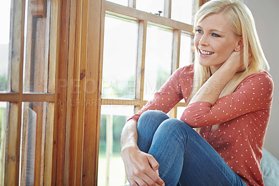 Buy stock photo A beautiful woman smiling and sitting near a window inside a house. Happy attractive blonde woman chilling at home. Happy female relaxing in an apartment enjoying the weekend and me time