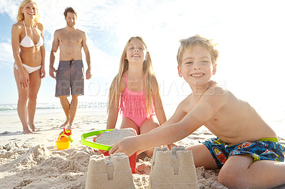Buy stock photo Shot of a young brother and sister building a sandcastle on the beach while their parents stand in the background