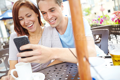 Buy stock photo A happy couple laughing at something on a cellphone while having coffee at a cafe