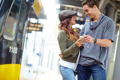 Buy stock photo A happy couple looking at each other and listening to music on a phone while waiting for a train with copyspace