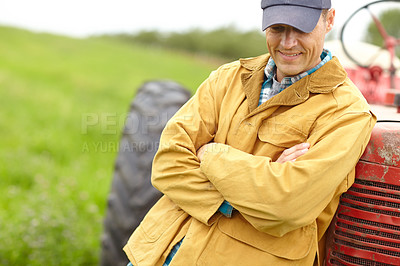 Buy stock photo Shot of a smiling farmer standing next to his tractor with his arms crossed and looking down