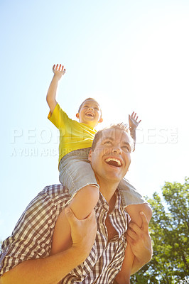 Buy stock photo Low angle shot of a cute young boy smiling in the sunshine and riding on his dad's shoulders