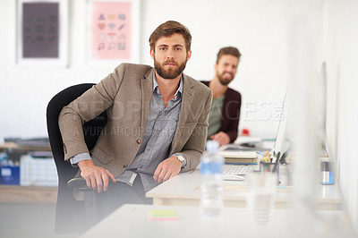 Buy stock photo Serious young businessman sitting at his desk with a colleague in the background
