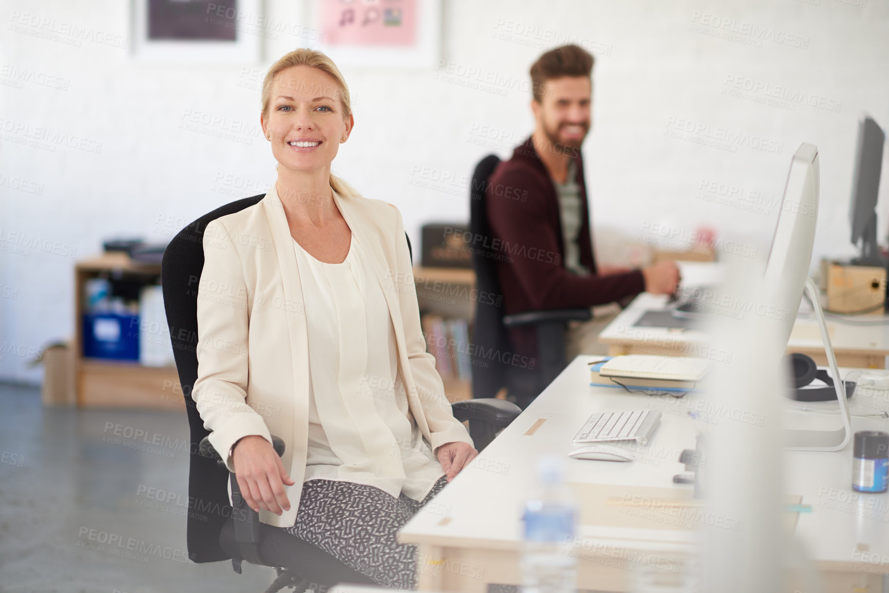 Buy stock photo Portrait of a smiling businesswoman sitting at her desk with her colleague in the background
