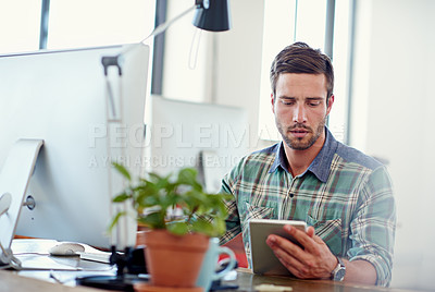 Buy stock photo Shot of a casually-dressed young man using a digital tablet at his desk