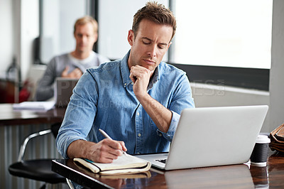Buy stock photo Shot of a man writing notes while working on a laptop in an office