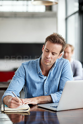 Buy stock photo Shot of a man writing notes while working on a laptop in an office