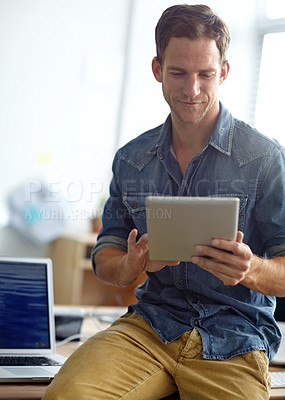 Buy stock photo A young man sitting on his desk at work while working on his digital tablet
