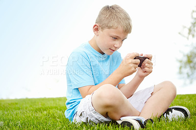 Buy stock photo A little boy engrossed on playing a game on his smartphone while lying outdoors