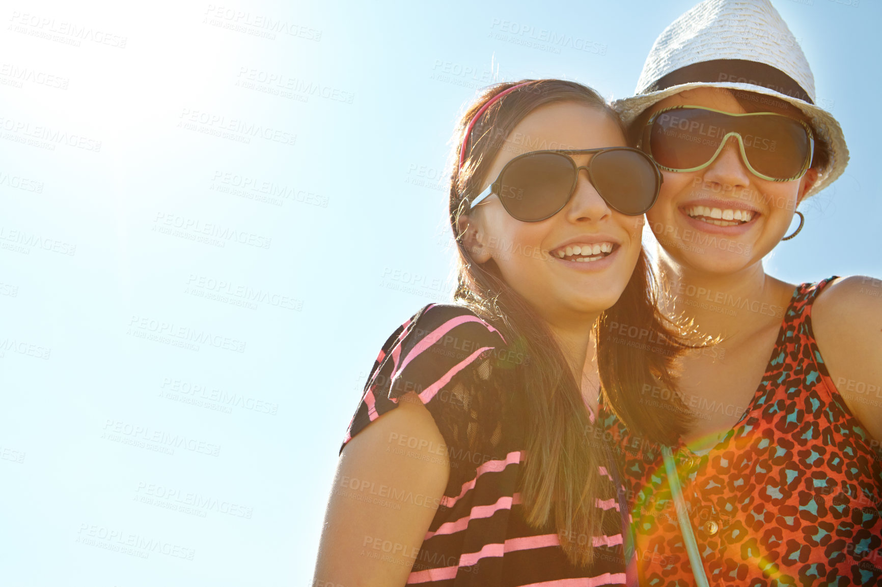 Buy stock photo Low angle view of two teenage girls standing with their arms around each other against a sunny sky