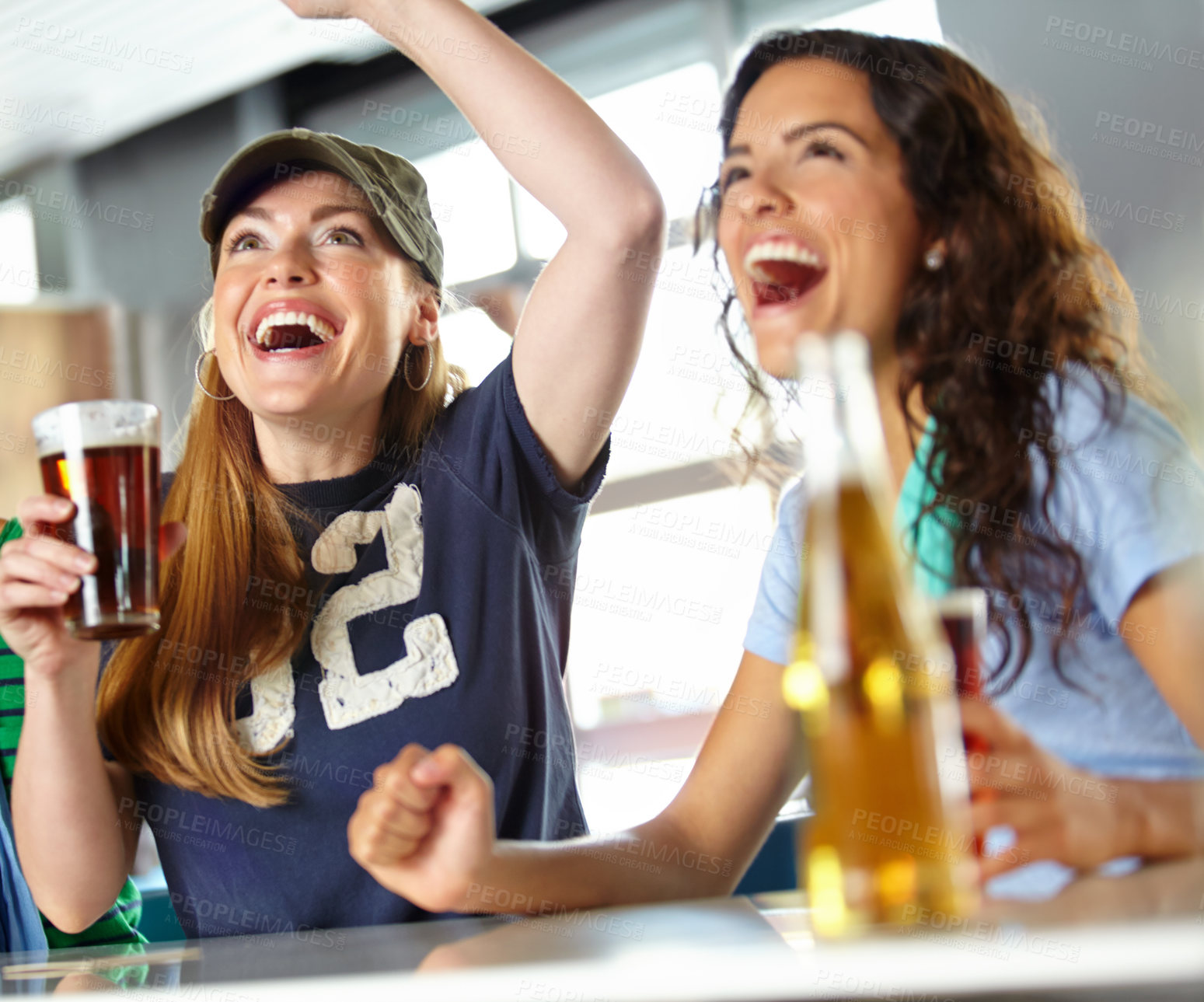 Buy stock photo Two girl friends cheering on their favourite team while kicking back with beers at the bar