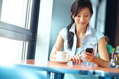 Buy stock photo A pretty asian woman reading a text message while enjoying a cup of coffee at a restaurant