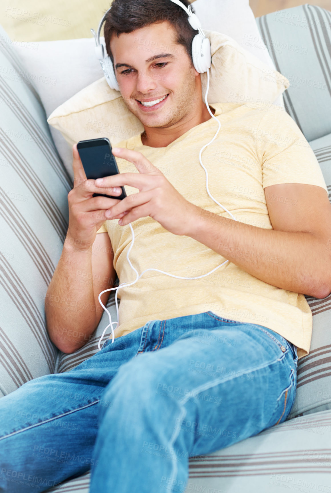 Buy stock photo Happy man, headphones and streaming with phone in relax on sofa for music, podcast or audio at home. Young male person with smile on mobile smartphone for online entertainment on couch at house