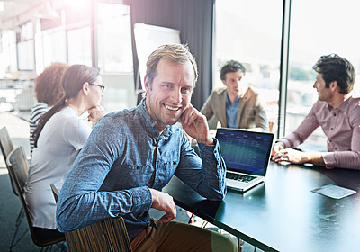 Buy stock photo Portrait of a smiling businessman sitting at a boardroom table with colleagues in the background