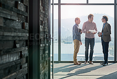 Buy stock photo Shot of male coworkers talking while standing in front of a window in an office