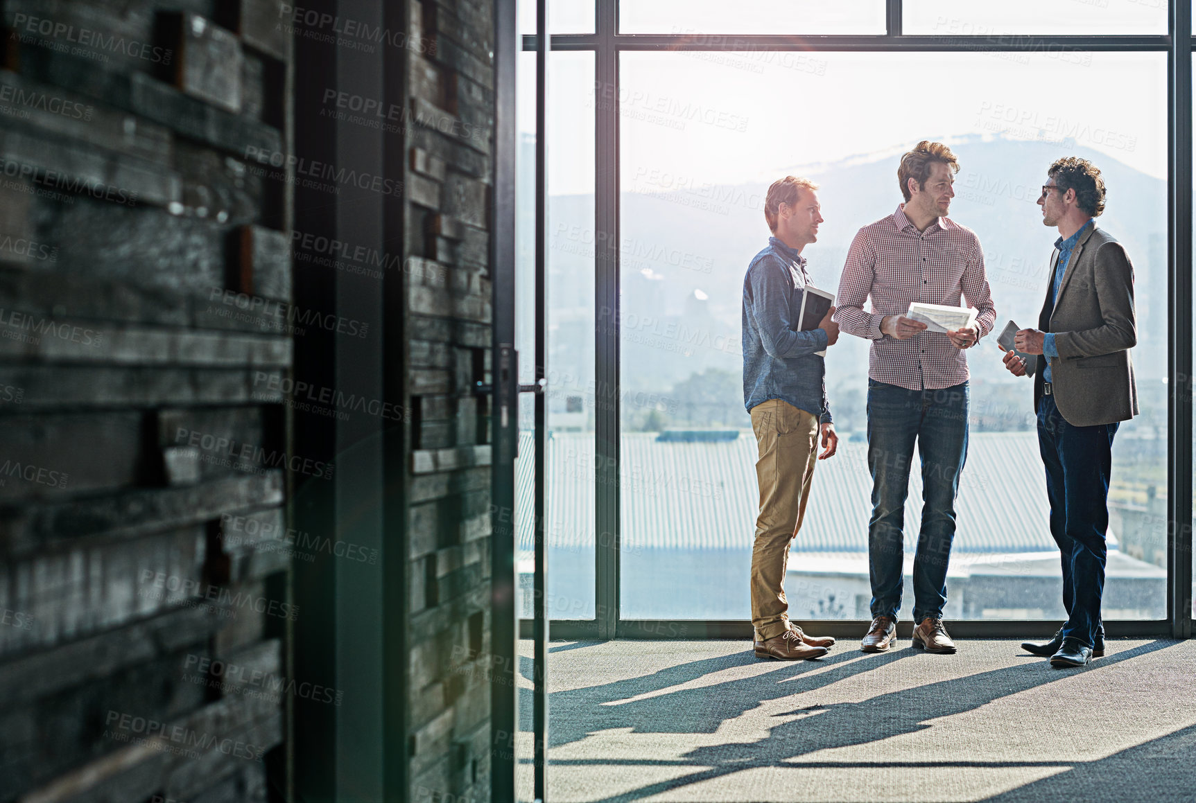 Buy stock photo Shot of male coworkers talking while standing in front of a window in an office