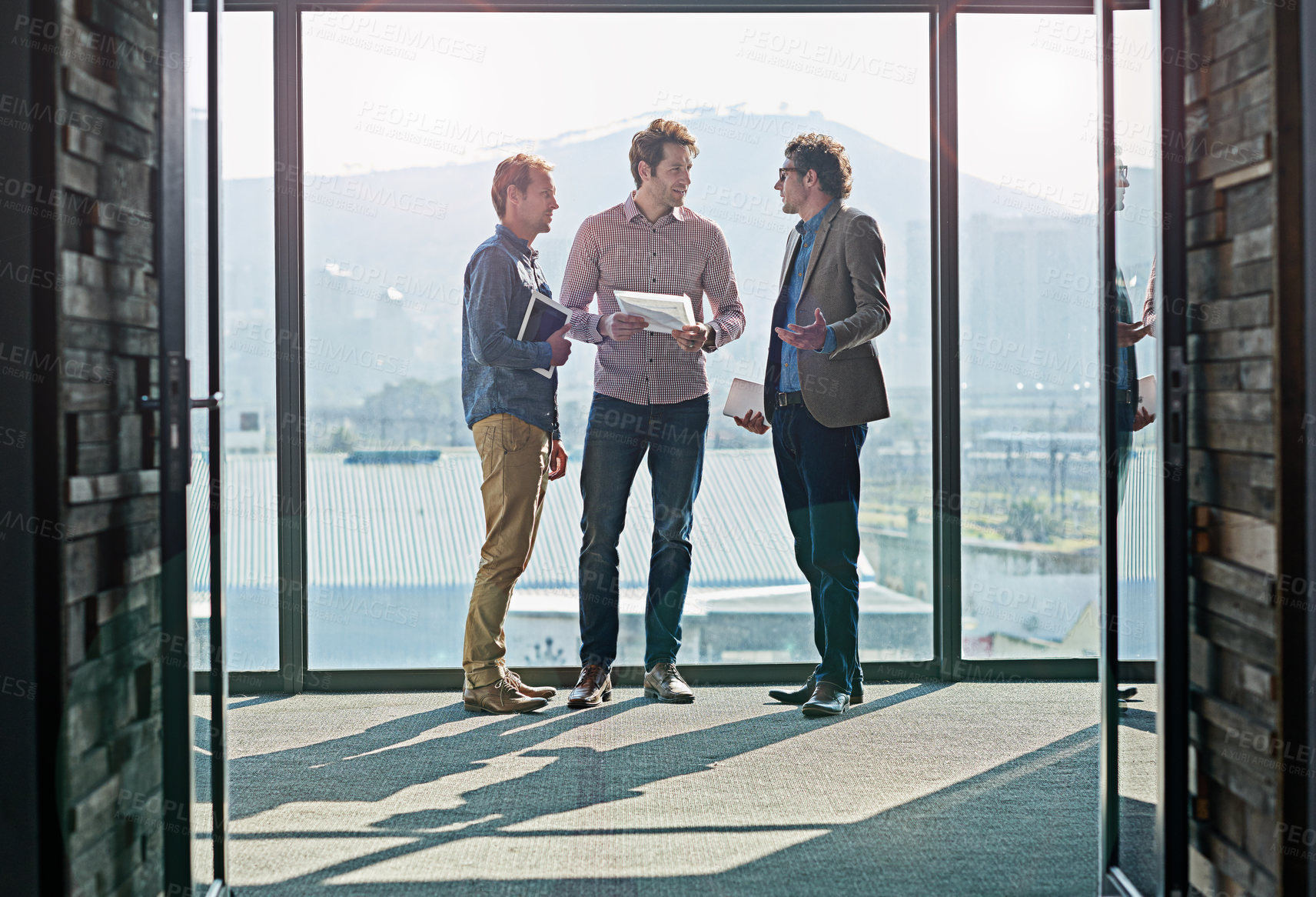 Buy stock photo Shot of male coworkers talking while standing in front of a window in an office