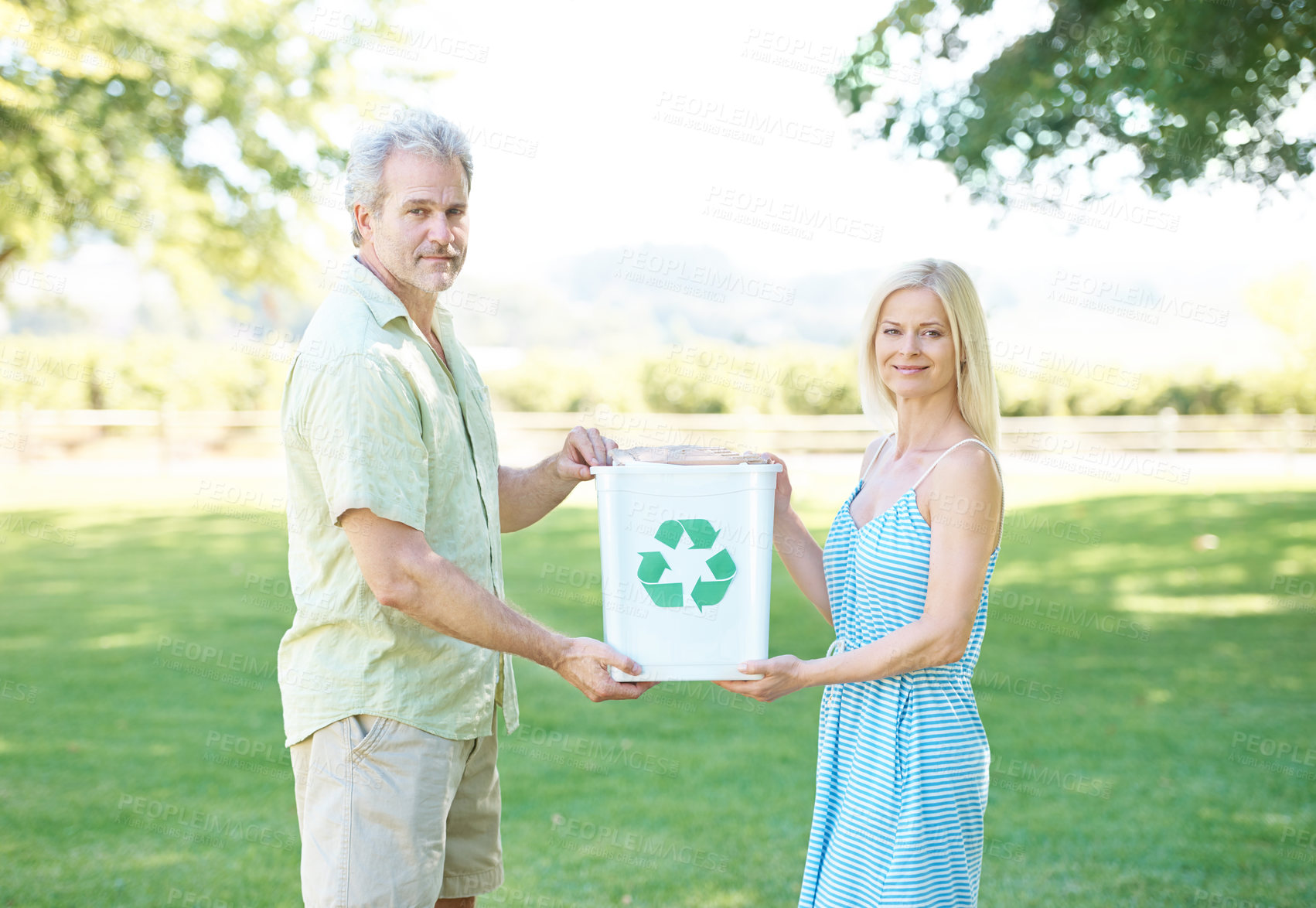 Buy stock photo A happy couple standing in a park holding a recycling bin and smiling at the camera