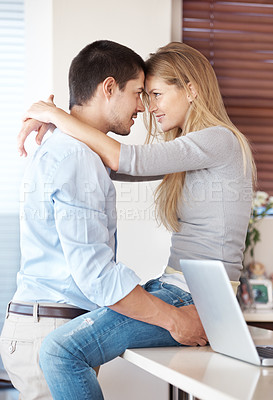 Buy stock photo A happy couple embracing in their kitchen with a laptop on the kitchen counter