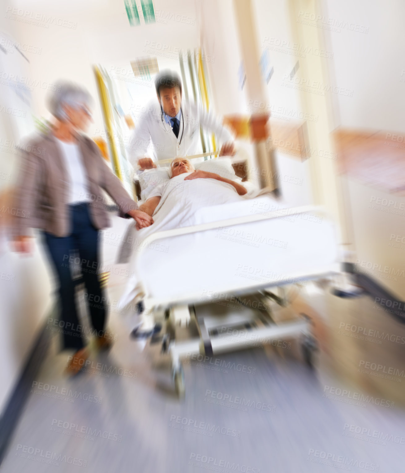 Buy stock photo Shot of a sick man on a bed being wheeled at speed through a hospital corridor with his wife at his side