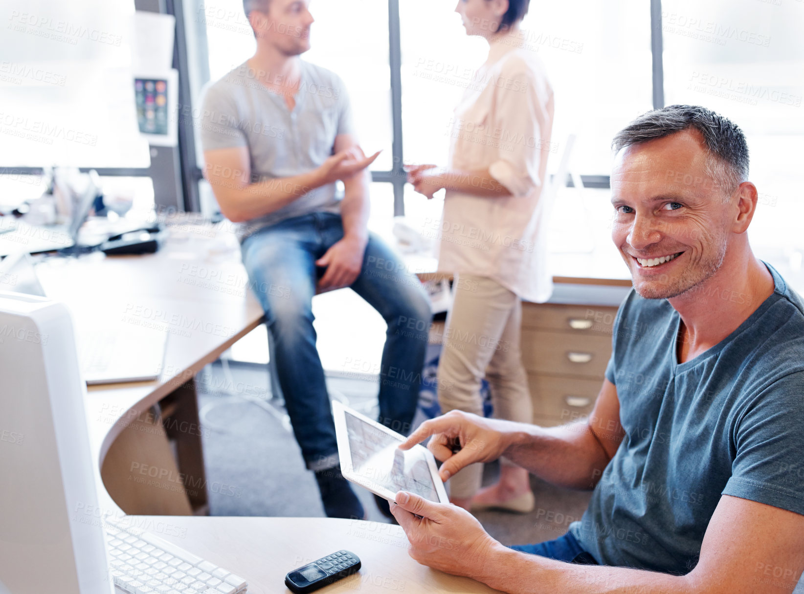 Buy stock photo Shot of an office worker using a tablet while his coworkers have a discussion in the background