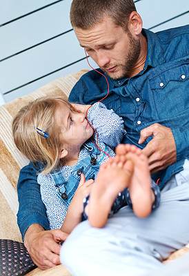 Buy stock photo Shot of an adorable little girl sitting on the sofa sharing headphones with her father