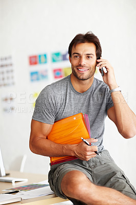 Buy stock photo A smiling handsome man sitting on an office desk and talking on his cellphone with folder under his arm and pen in hand