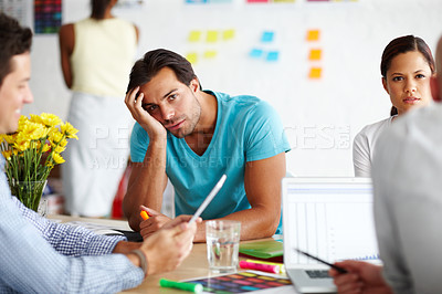 Buy stock photo Portrait of a bored looking man sitting at an office table with head rested on his hand and surrounded by colleges 
