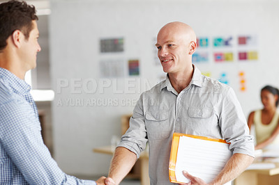 Buy stock photo Shot of two young men shaking hands in an office with a colleague working at a desk in the background